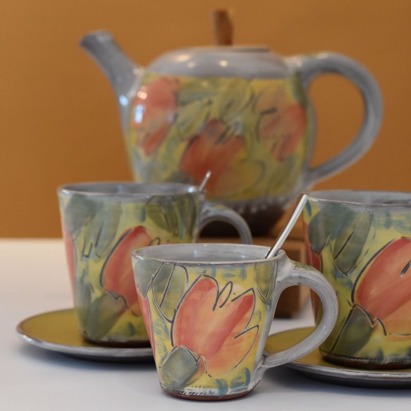 handgetoepferte-keramik-tasse-konisch-klein-gross-bluete-gelb-1 Handgetöpferte Keramiktasse mit Löffel und weiteren Keramiktassen und einer Teekanne aus Keramik im Hintergrund.